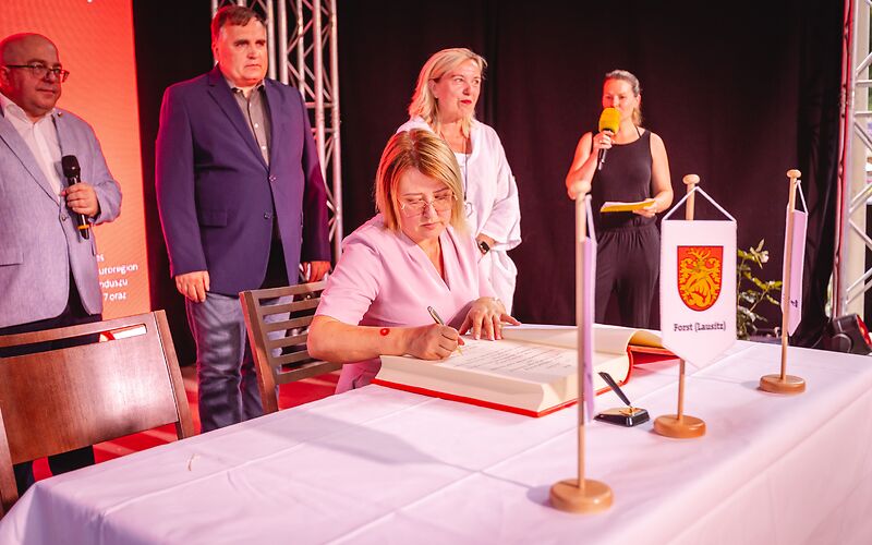 Frau Beata Kowalczuk, Bürgermeisterin von Brody, sitzt an einem mit einem weißen Tischtuch gedeckten Tisch, auf dem das goldene Buch der Stadt Forst (Lausitz) aufgeschlagen liegt. Hinter ihr stehen u. a. der Vorsitzende der Stadtverordnetenversammlung Ingo Paeschke und die Bürgermeisterin von Forst Simone Taubenek.