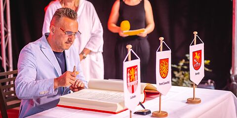 Herr Janusz Dudojć (Bürgermeister von Lubsko) sitzt an einem mit einem weißen Tischtuch gedeckten Tisch, auf dem das goldene Buch der Stadt Forst (Lausitz) aufgeschlagen liegt. Vorn auf dem Tisch stehen die Wappen der Städte Forst (Lausitz), Lubsko und Brody.