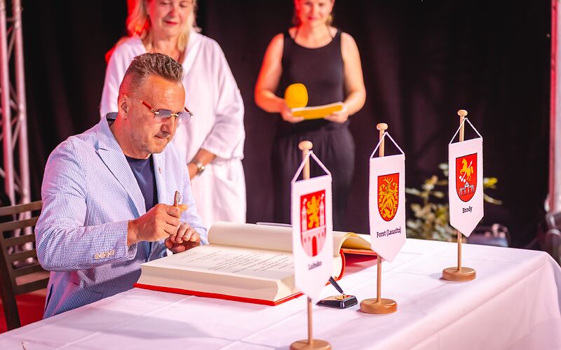 Herr Janusz Dudojć (Bürgermeister von Lubsko) sitzt an einem mit einem weißen Tischtuch gedeckten Tisch, auf dem das goldene Buch der Stadt Forst (Lausitz) aufgeschlagen liegt. Vorn auf dem Tisch stehen die Wappen der Städte Forst (Lausitz), Lubsko und Brody.