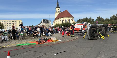 Eine Vorführung durch den Rettungsdienst: Ein Auto liegt seitlich auf dem Platz, Männer in Uniform und ein Rettungswagen stehen in unmittelbarer Nähe. Viele Zuschauer stehen um sie herum.