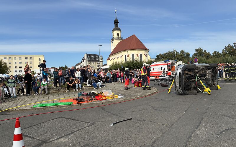 Eine Vorführung durch den Rettungsdienst: Ein Auto liegt seitlich auf dem Platz, Männer in Uniform und ein Rettungswagen stehen in unmittelbarer Nähe. Viele Zuschauer stehen um sie herum.