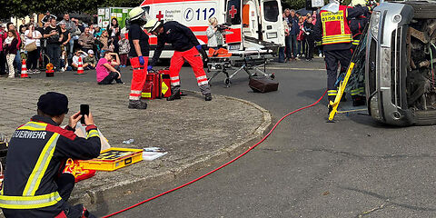 Vorführung vor einem Publikum: Zwei Rettungskräfte stehen bei einem auf der Seite liegenden Auto.