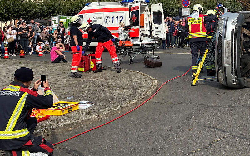 Vorführung vor einem Publikum: Zwei Rettungskräfte stehen bei einem auf der Seite liegenden Auto.