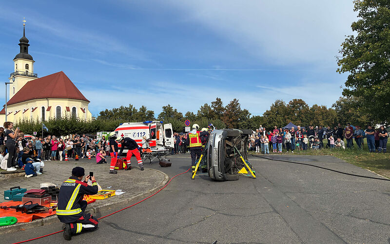 Vorführung vor einem Publikum: Zwei Rettungskräfte stehen bei einem auf der Seite liegenden Auto.