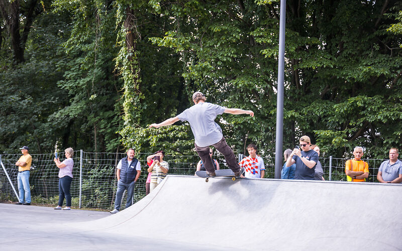 Skateboardfahrer führt Trick auf einer Rampe aus, im Hintergrund mehrere zuschauende Personen vor Bäumen