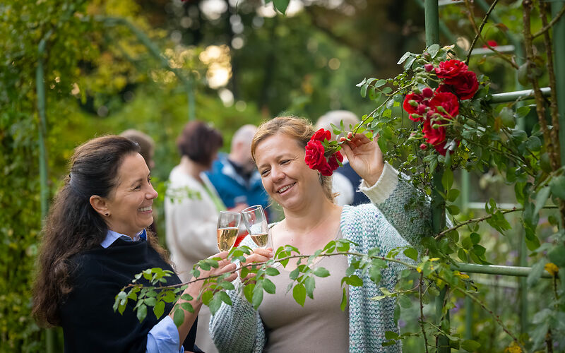 zwei Frauen stehen an einer Rosenranke mit roten Rosenblüten und stoßen mit zwei Gläsern an