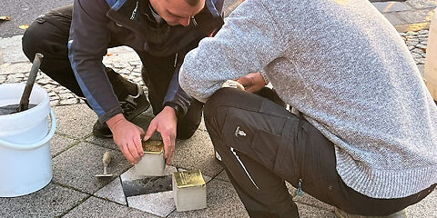 Zwei Mitarbeiter des Betriebsamtes bei der Verlegung der Stolpersteine. Sie hocken und die Steine liegen direkt neben dem Loch.