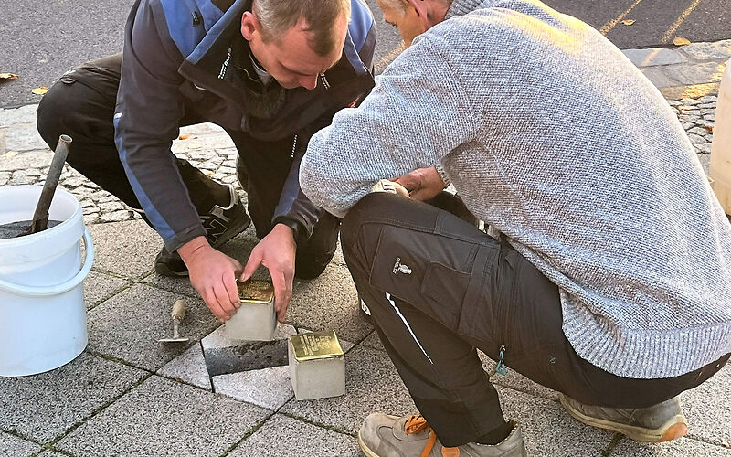 Zwei Mitarbeiter des Betriebsamtes bei der Verlegung der Stolpersteine. Sie hocken und die Steine liegen direkt neben dem Loch.