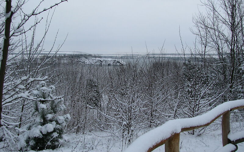 Blick auf den Klinger See von einem Weg aus, der durch einen mit Schnee bedeckten Holzzaun eingefasst ist. Vor dem See stehen verschneite kahle Bäume. In den See hinein ragt eine Landzunge.