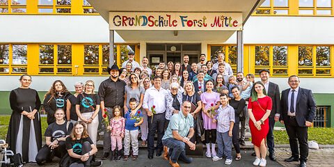 Gruppenfoto der Mitglieder der Familie Levy, der Teilnehmenden aus der Verwaltung und der Grundschule Forst Mitte, sowie weitere Teilnehmer auf den Stufen am Haupteingang der Grundschule Forst Mitte.