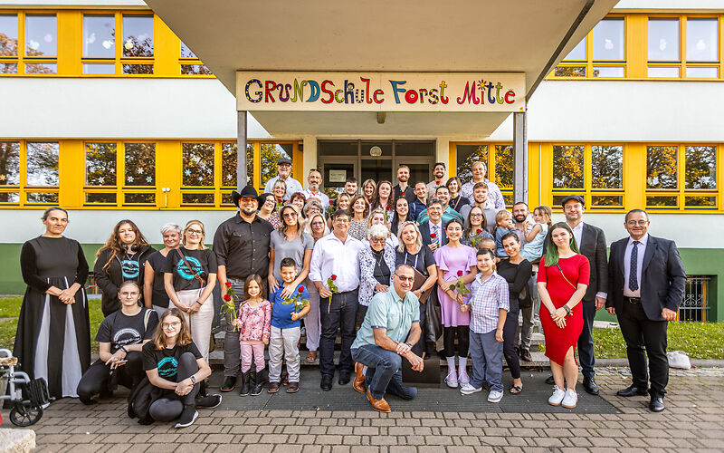 Gruppenfoto der Mitglieder der Familie Levy, der Teilnehmenden aus der Verwaltung und der Grundschule Forst Mitte, sowie weitere Teilnehmer auf den Stufen am Haupteingang der Grundschule Forst Mitte.