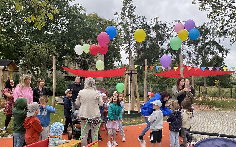 Kinder der Kita und Gäste auf der Einweihungsfeier. Es gibt Luftballons und eine Girlande als Deko. 