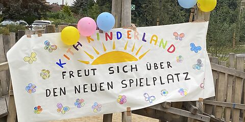 Ein mit bunten Blumen und Schmetterlingen gestaltetes Plakat auf dem "Kita Kinderland freut sich über den neuen Spielplatz" steht.