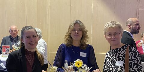 Drei lächelnde Frauen an einem der Stehtische, auf dem Stehtisch sind Blumen und Gläse mit Getränken und Salaten.