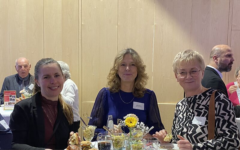 Drei lächelnde Frauen an einem der Stehtische, auf dem Stehtisch sind Blumen und Gläse mit Getränken und Salaten.