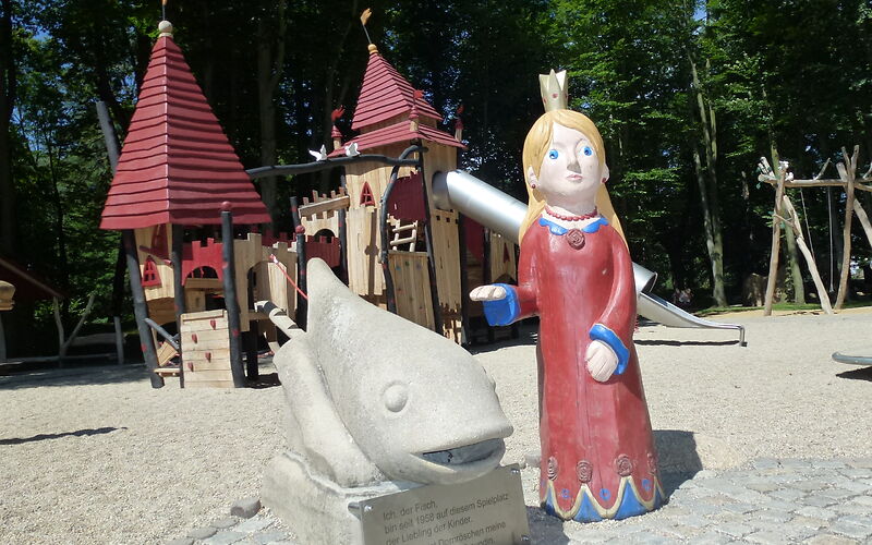 Eine Holzfigur einer prizessin mit blonden Haaren, blauen Augen und einem roten Kleid steht neben einer Statue eines Fisches. Im Hintergrund ist ein Klettergerüst, welches wie ein Schloss aussieht auf einem Sandplatz. 