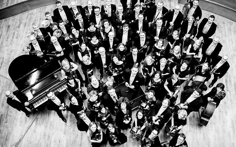 ein schwarz-weiß Bild zeigt ein Orchester der Zuschauer guckt von oben auf die Gruppe runter, die Muiker lächeln nach oben