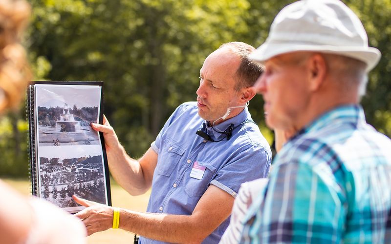 In der Mitte zeigt der Gästeführer mit blauen Hemd eine Mappe mit einer historischen Ansicht des Kasakenbunnen, eine Frau und ein Herr mit Hut in Rückenansicht betrachten das Bild