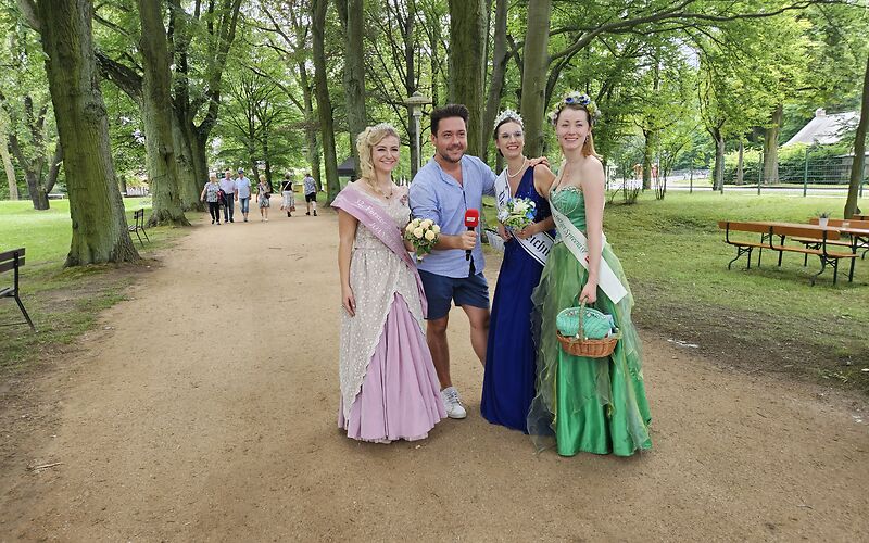 Auf einem Weg im Rosenkarten, an welchem links und rechts Bäume stehen, steht die Forster Rosenkönigin zusammen mit einem Roporter vom rbb, der Peitzer Teichnixe und der Spremberger Spreenixe.