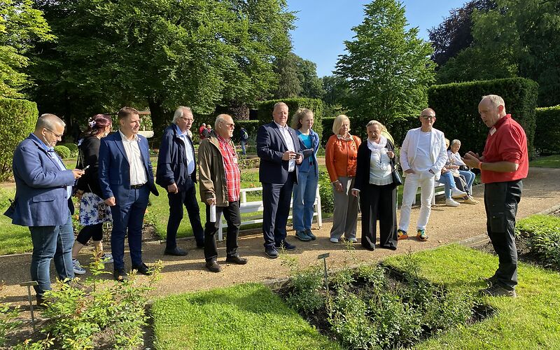Die Gruppe steht auf einem Sandweg, an dem zu beiden Seiten auf einer Wiese quardratische Rosenbeete angelegt sind. Die Sonne scheint. 
