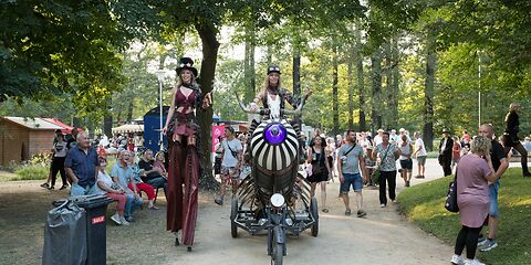 Eine Stelzenläuferin und ein Steampunk-Dreirad mit Radkete, auf der die Fahrerin sitzt.