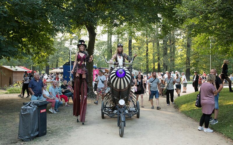 Eine Stelzenläuferin und ein Steampunk-Dreirad mit Radkete, auf der die Fahrerin sitzt.