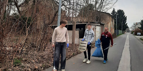 3 Personen gehen eine Staße entlang. Sie tragen eine Greifzange, einen blauen Müllsack und einen dreckigen Sitz eines Stuhles.