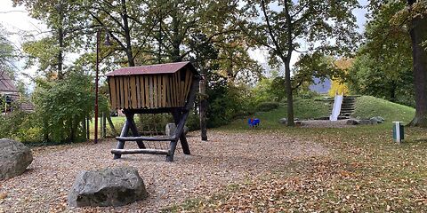 Auf einer Sandfläche steht ein Holzklettergerüst, der obere Teil ist ein Haus auf dicken Balken, dass über eine Kletternetz erreicht werden kann. Im Hintergrund steht am Rand der Fläche eine blaue Federwippe.