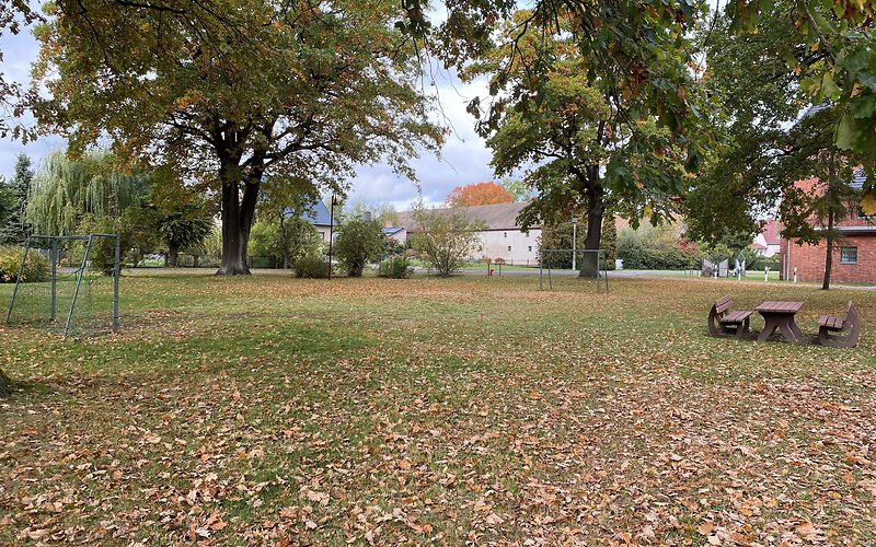 Auf der Wiese des Spielplatzes sind 2 Fußballtore gegenüber aufgestellt. Rechts stehen 2 Holzbänke und eine Holztisch. Auf der Wiese liegen überall Blätter. Im Hintergrund stehen mehrere Bäume und Sträucher.