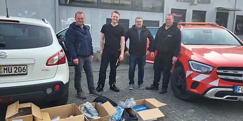4 Männer in dunkler Kleidung auf einem Parkplatz. Vor Ihnen stehen Pappkisten mit Untensilien, z. B. blaue Müllsäcke.