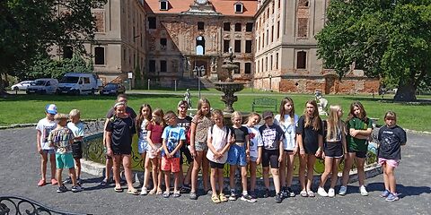 Die Schülerinnen und Schüler stehen in einer Reihe vor einem Brunnen. Im Hintergrund befindet sich die Schlossruine des Schlosses in Brody. Das Schloss ist ein U-Förmiges Gebäude mit einer großen Uhr in der Mitte. Es steht auf einer Wiese, links und rechts befinden sich große Bäume.