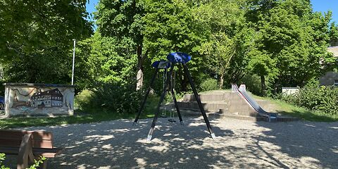 Die Metallrutsche befindet sich auf einem Hügel rechts hinten im Bild. Vorn ist eine Bank von hinten zu sehen. Mittig steht die Doppelschaukel im Schattend er Bäume.