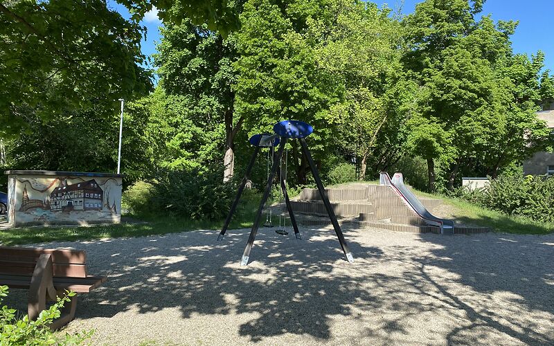 Die Metallrutsche befindet sich auf einem Hügel rechts hinten im Bild. Vorn ist eine Bank von hinten zu sehen. Mittig steht die Doppelschaukel im Schattend er Bäume.
