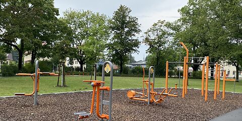 Sportgeräte auf dem Spielplatz, gestaltet in den farben orange und grau sin im Vodergrund, darunter Arm-Bauchmuskeltrainer und Crisstrainer.