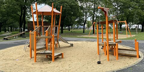 Auf einem Sandplatz steht in Turm mit Geüst mit Dach aus Orangenen Stangen und einem Aufstieg aus Holz. An Turm ist eine silberne Rutsche.