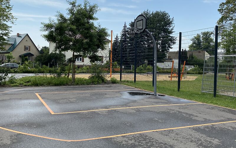 Vor einem Zaun ist das Feld um einen Basketballkorb auf Asphaltboden mit orangenen Linien aufgezeichnet. 