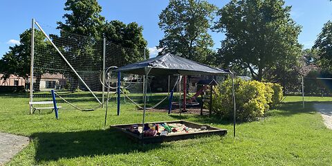 Ein überdachter Sandkasten, auf einer Wiese. Im Sandkasten liegt Sandspielzeug.