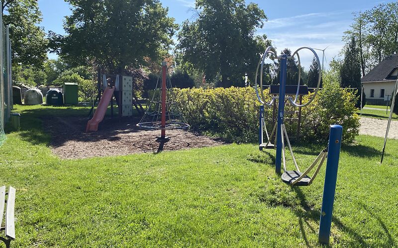 Eine Rutsche mit der Rutschbahn nach vor, ein Seilbaum auf eier Sandfläche und der Doppelschwinger auf der Wiese
