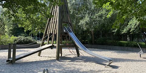 Ein Kletterturm mit Rutsche steht zentral auf einem Sandplatz. Vorn im Bild steht eine Federwippe für 4 Personen. Im Hintergrund sind Bäume und Wiese.