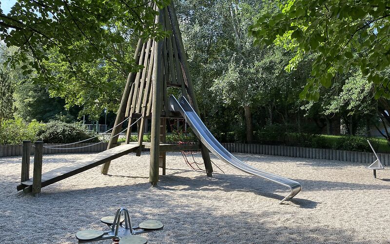 Ein Kletterturm mit Rutsche steht zentral auf einem Sandplatz. Vorn im Bild steht eine Federwippe für 4 Personen. Im Hintergrund sind Bäume und Wiese.