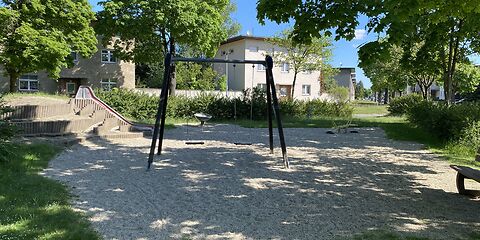 Eine Doppelschaukel steht auf einer Sandfläche, dahinter befindet sich eine Rutsche. Die Fläche ist umgeben von Wiese und Bäumen, im Hintergrund steht ein Haus, die sonne scheint.