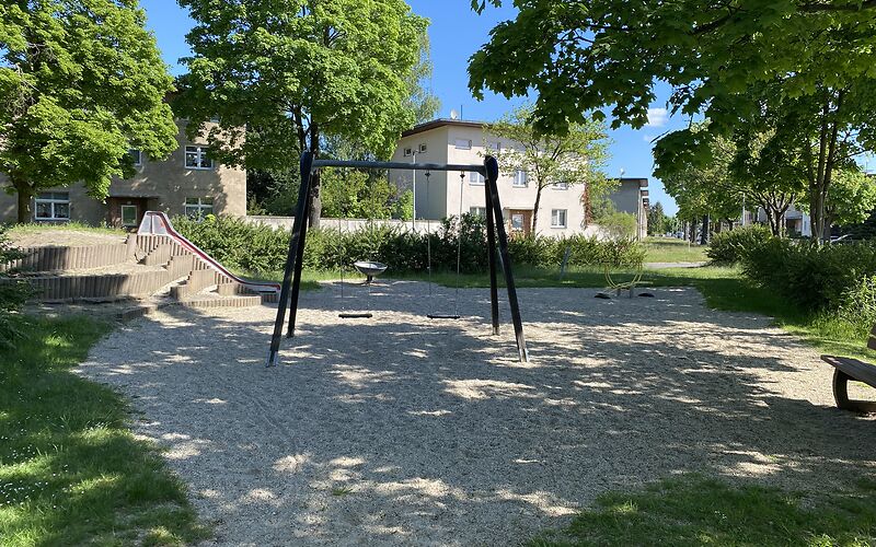 Eine Doppelschaukel steht auf einer Sandfläche, dahinter befindet sich eine Rutsche. Die Fläche ist umgeben von Wiese und Bäumen, im Hintergrund steht ein Haus, die sonne scheint.