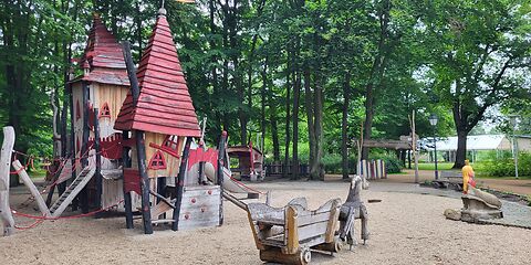 Ein Klettergerüst mit zwei Roten Türmen aus Holz, welches auf einem Sandplatz steht. Davor steht ein Holzwagen mit einem Holzpferd davor. Im Hintergrund befinden Bäume und der Eingang zum Spielplatz. 