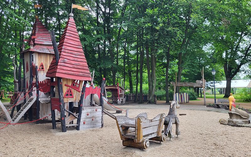 Ein Klettergerüst mit zwei Roten Türmen aus Holz, welches auf einem Sandplatz steht. Davor steht ein Holzwagen mit einem Holzpferd davor. Im Hintergrund befinden Bäume und der Eingang zum Spielplatz. 