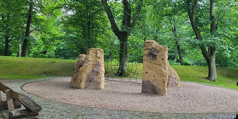 Ein Kletternetz aus Seilen, welches zwischen zwei Felsen gespannt ist. Um das Gerüst herum ist eine runde Fläche aus Sand, dahinter befinden sich Wiese und Bäume. Im Vorderung ist gepflastert.