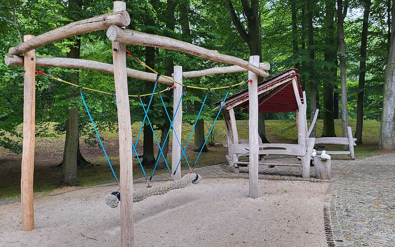 An einem Holzgestellt aus runden Balken hängt ein dickes Seil zum Klettern über einer Sandfläche. Auf der linken Seite stehen Bäumen, rechts ist ein geflasterte weg. Im Hintergrund stehen 2 Holzbämke sich gegenüber.