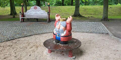 Eine König (Holzfigur) steht auf einer Wederwippen auf einem Sandplatz. Im Hintergrund ist ein gepflanster Weg mit einer Bank und Bäume  auf einer Wiese.