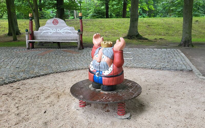 Eine König (Holzfigur) steht auf einer Wederwippen auf einem Sandplatz. Im Hintergrund ist ein gepflanster Weg mit einer Bank und Bäume  auf einer Wiese.