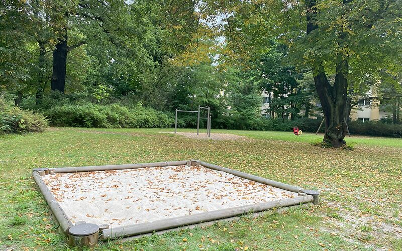 Ein Sandkasten mit Holzumrandung auf einer Wiese. Im Sand und auf der Wiese liegen bunte Blätter.