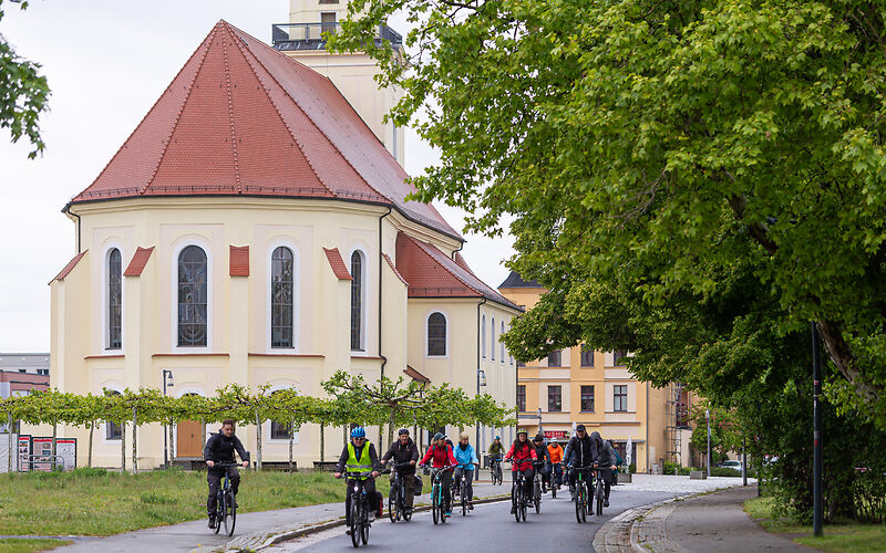 Die Gruppe fährt mit Fahrrädern auf einer Straße, im Hintergrund links sieht man den Platanenhain und die Stadtkirche.